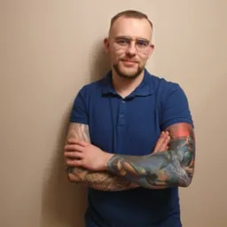 Mężczyzna w okularach i niebieskiej koszulce polo z tatuażami na obu ramionach, stojący na tle beżowej ściany.