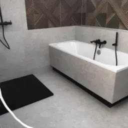 Nowoczesna łazienka z szarymi płytkami, białą wanną z czarną armaturą i dekoracyjnym panelem ściennym z geometrycznym wzorem nad wanną.
