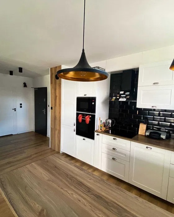 Nowoczesna kuchnia w Katowicach z białymi szafkami, czarnym piekarnikiem i płytkami w stylu metro, oświetlona wiszącą lampą z elementami w kolorze złota.