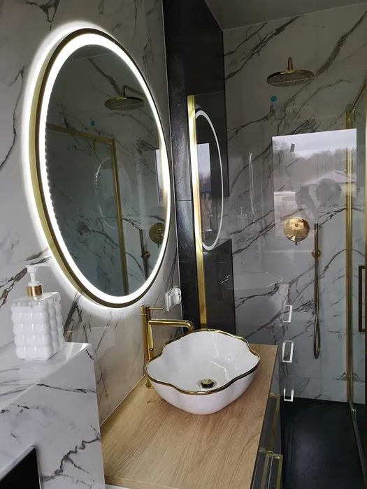 Nowoczesna łazienka z marmurowymi płytkami, okrągłym lustrem LED, złotą armaturą i umywalką w kształcie kwiatu.