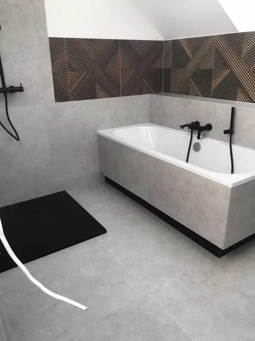 Nowoczesna łazienka z szarymi płytkami, białą wanną z czarną armaturą i dekoracyjnym panelem ściennym z geometrycznym wzorem nad wanną.