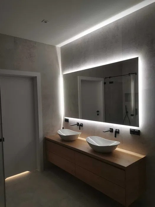 Nowoczesna łazienka z dwoma owalnymi umywalkami na drewnianej szafce, podświetlane lustro LED na szarej ścianie z płytek, widoczny fragment drzwi.