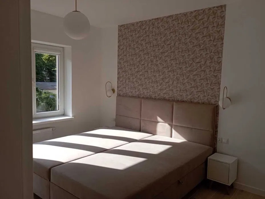 Jasna sypialnia z tapetą w geometryczne wzory, łóżko kontynentalne z wezgłowiem w kolorze pudrowego różu, okno z widokiem na zieleń i lampy ścienne w kształcie owali.