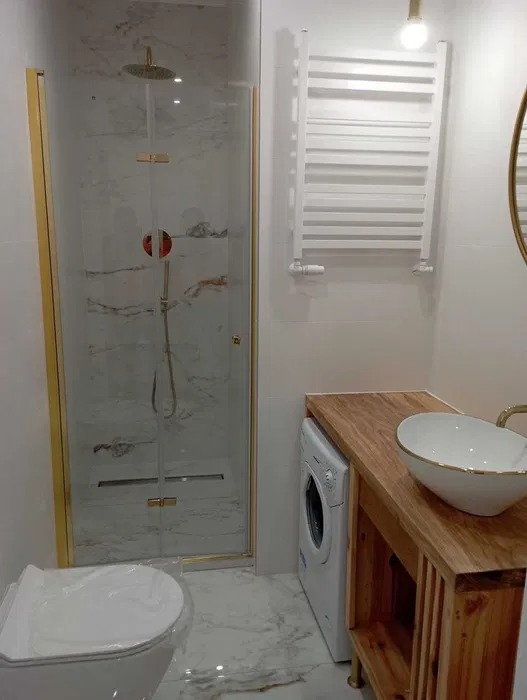 Nowoczesna łazienka z marmurowymi płytkami, kabiną prysznicową ze złotymi ramami, umywalką na drewnianym blacie i pralką schowaną pod blatem.