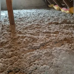 Ocieplenie poddasza wełną mineralną, widoczna drewniana konstrukcja dachu i fragment okna. Szara, włóknista izolacja pokrywa całą powierzchnię.