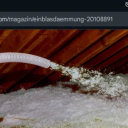 Aplikacja izolacji w przestrzeni poddasza za pomocą węża i dmuchawy. Widoczne drewniane krokwie i biała izolacja. Osoba w żółtych rękawicach obsługuje urządzenie.