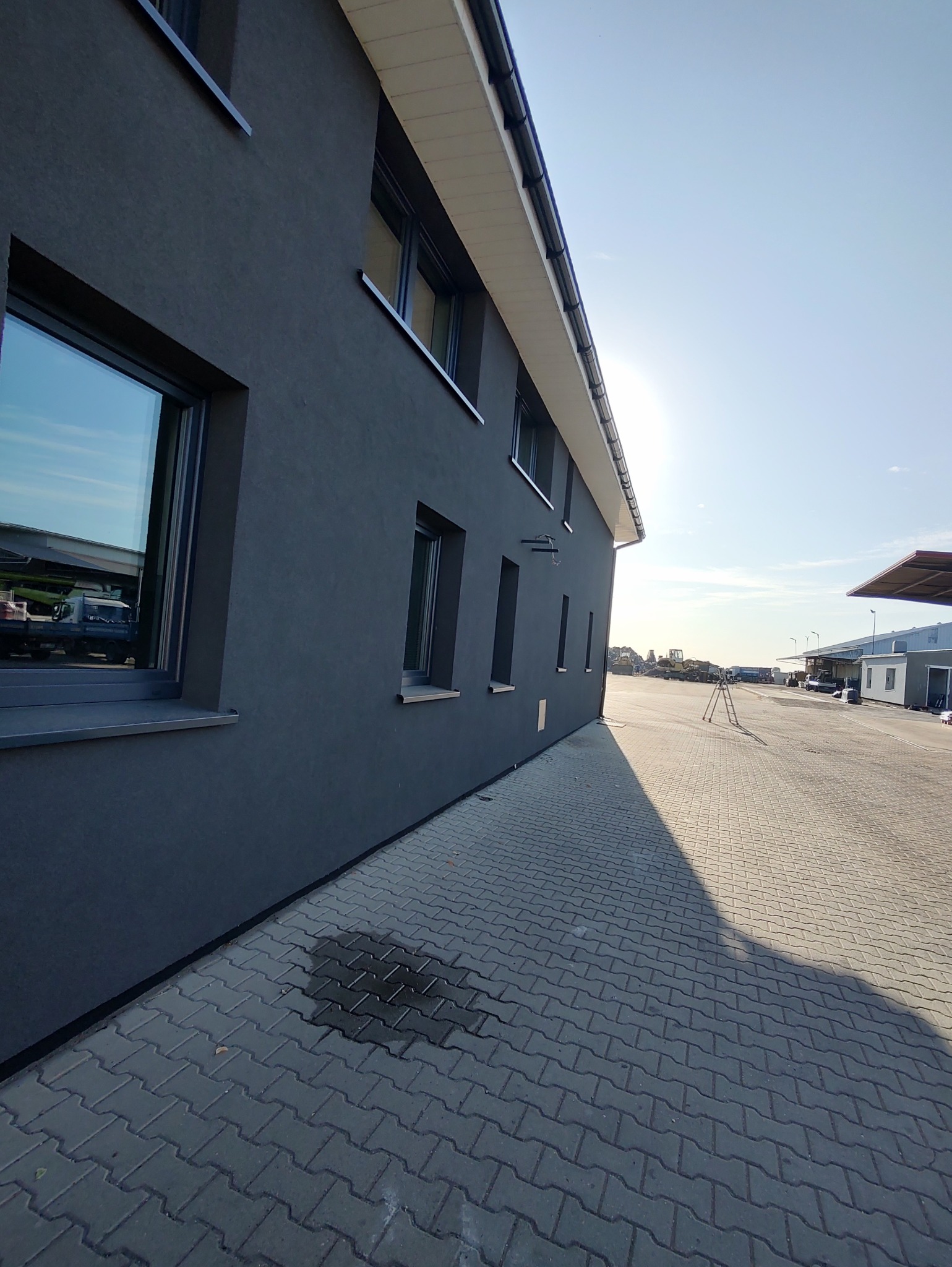 Elewacja budynku w kolorze grafitowym z widocznymi oknami i kostką brukową na zewnątrz, oświetlona słońcem. Widok na zewnątrz budynku.