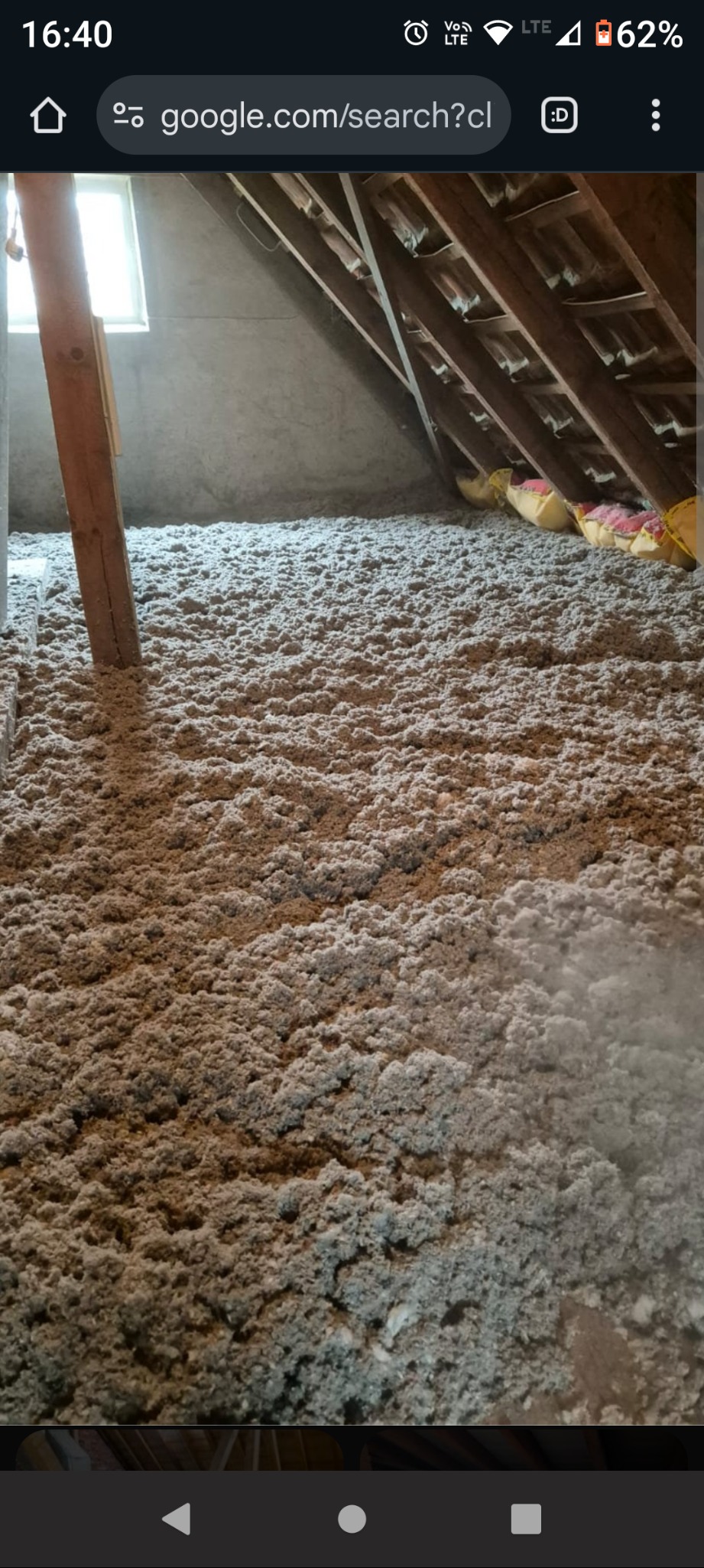 Ocieplenie poddasza wełną mineralną, widoczna drewniana konstrukcja dachu i fragment okna. Szara, włóknista izolacja pokrywa całą powierzchnię.