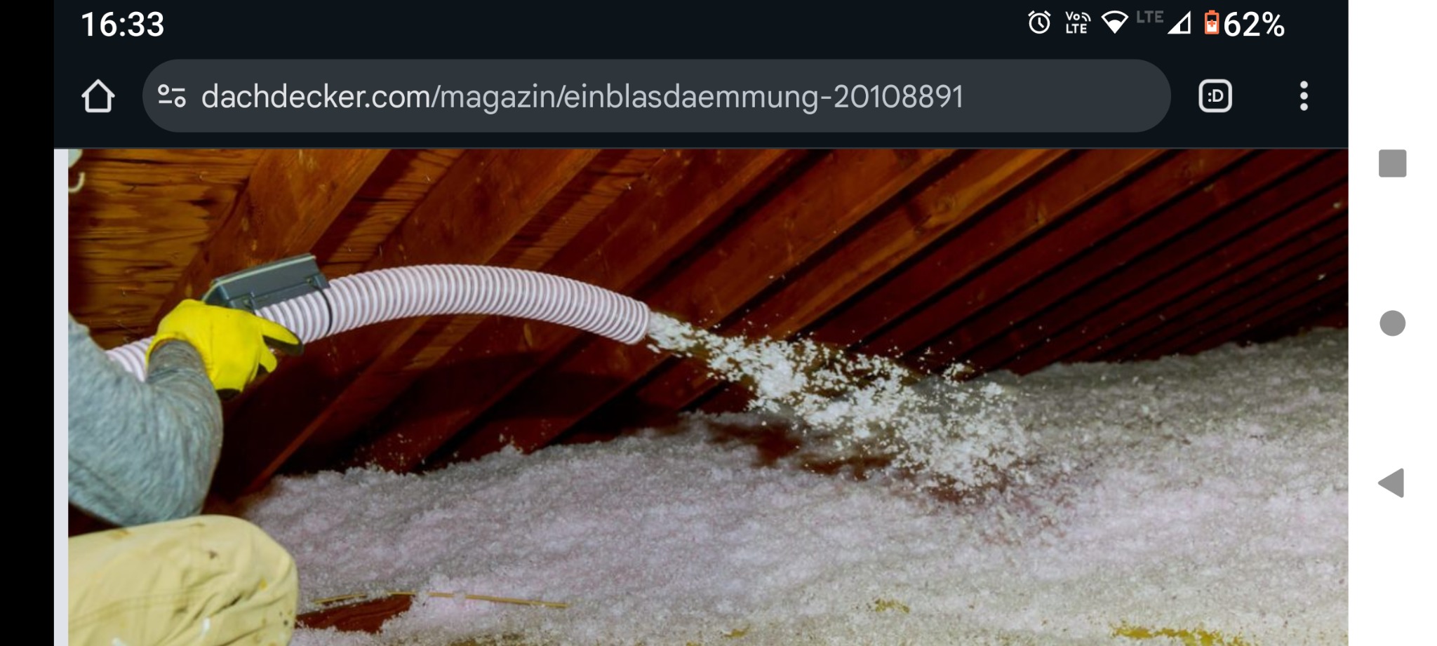 Aplikacja izolacji w przestrzeni poddasza za pomocą węża i dmuchawy. Widoczne drewniane krokwie i biała izolacja. Osoba w żółtych rękawicach obsługuje urządzenie.