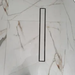 Białe płytki imitujące marmur z odpływem liniowym w kabinie prysznicowej, widoczny fragment roboczego buta.