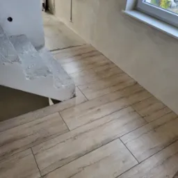 Ułożone panele podłogowe imitujące drewno, prowadzące od schodów w surowym stanie do okna z widokiem na zieleń.