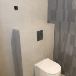 Wnętrze łazienki z podwieszaną toaletą, beżowe płytki ścienne i podłogowe, szare płytki dekoracyjne w kształcie rombów na ścianie, widoczna instalacja spłuczki z chromowanym przyciskiem, otwór...