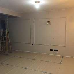 KTM-REM - Pokój w trakcie prac wykończeniowych: ściany pomalowane na szaro z dekoracyjnymi listwami, zabezpieczona podłoga, drabina malarska w rogu, lampa ścienna w stylu retro.