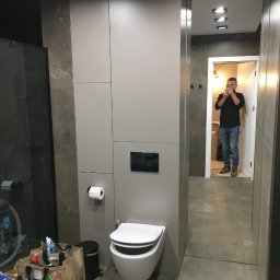 KTM-REM - Nowoczesna łazienka z szarymi płytkami imitującymi marmur, podwieszana toaleta z czarnym przyciskiem spłukującym, widoczny fragment kabiny prysznicowej i odbicie osoby robiącej zdjęcie w lustrze.