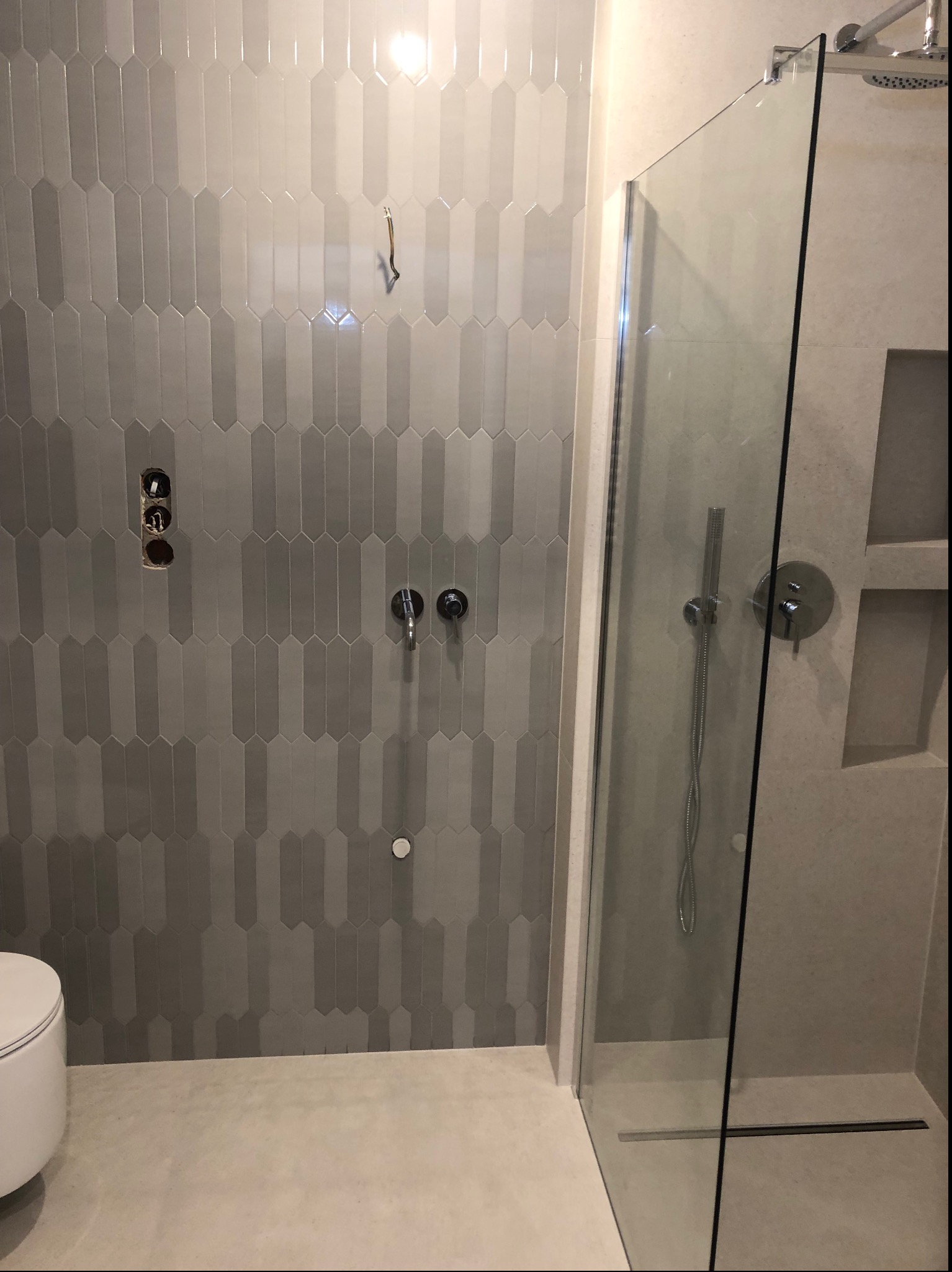 Szara łazienka w trakcie wykańczania z kabiną prysznicową typu walk-in, pionowo ułożone płytki w kształcie wydłużonych ośmiokątów, widoczne przyłącza do baterii i wystające przewody elektryczne.