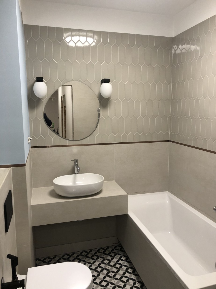 Jasna łazienka z geometrycznymi płytkami na ścianie, okrągłym lustrem z dwoma kulistymi lampami, białą umywalką na betonowym blacie i czarno-białą podłogą w marokański wzór.
