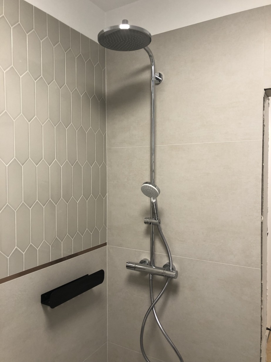 Wykończona łazienka z chromowaną baterią prysznicową, szarą mozaiką w kształcie rombów i czarną półką na akcesoria.