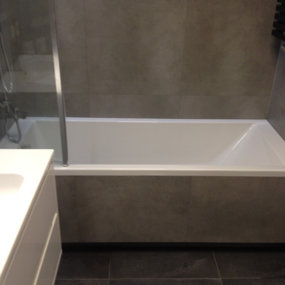 Nowoczesna łazienka z wanną obudowaną szarymi płytkami imitującymi beton, kabiną prysznicową ze szklanym panelem i umywalką z białym blatem.