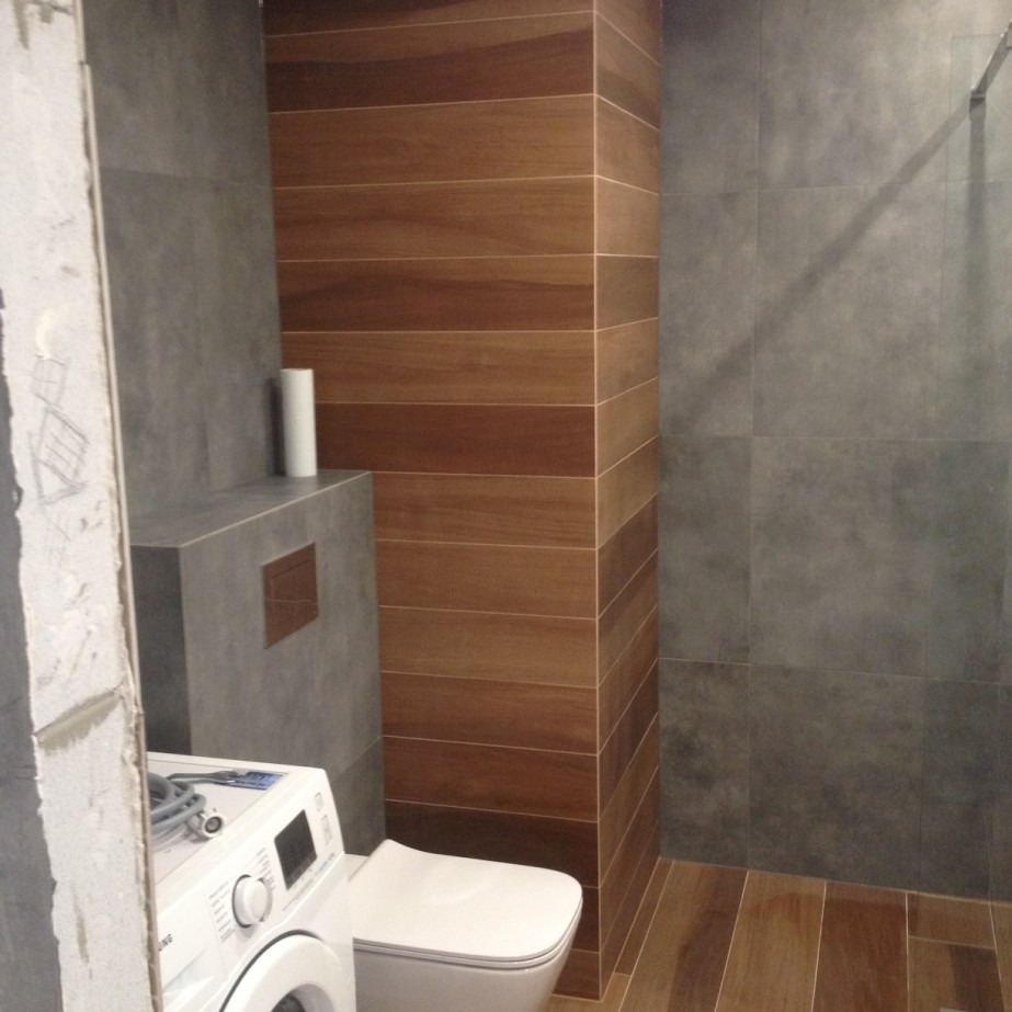 Nowoczesna łazienka z szarymi płytkami imitującymi beton i drewnopodobnymi panelami na kolumnie, widoczna pralka i toaleta.