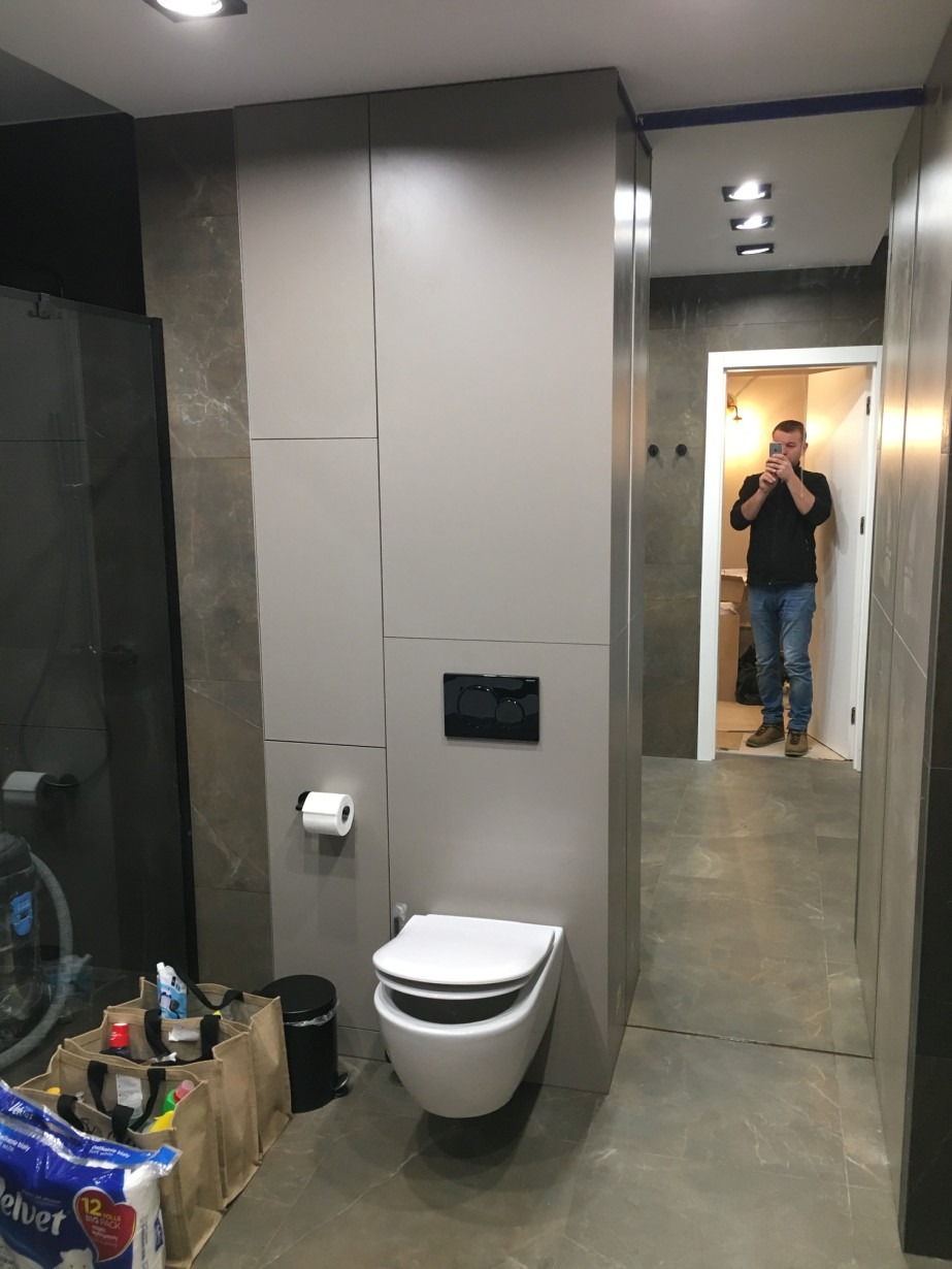 Nowoczesna łazienka z szarymi płytkami imitującymi marmur, podwieszana toaleta z czarnym przyciskiem spłukującym, widoczny fragment kabiny prysznicowej i odbicie osoby robiącej zdjęcie w lustrze.