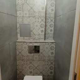 Kompaktowa toaleta podwieszana z szarą mozaikową ścianą, chromowany przycisk spłukiwania i rewizja w zabudowie.