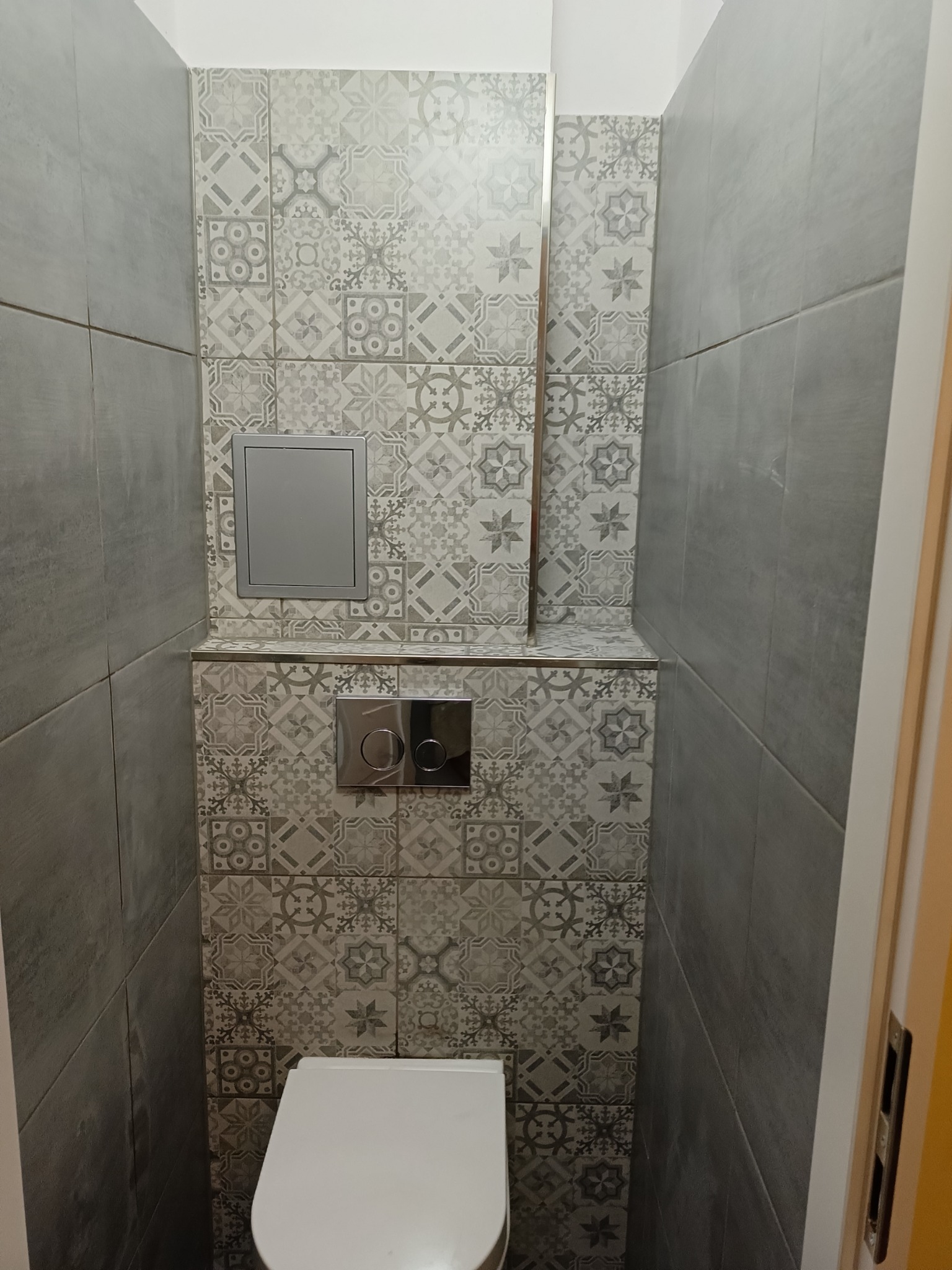 Kompaktowa toaleta podwieszana z szarą mozaikową ścianą, chromowany przycisk spłukiwania i rewizja w zabudowie.