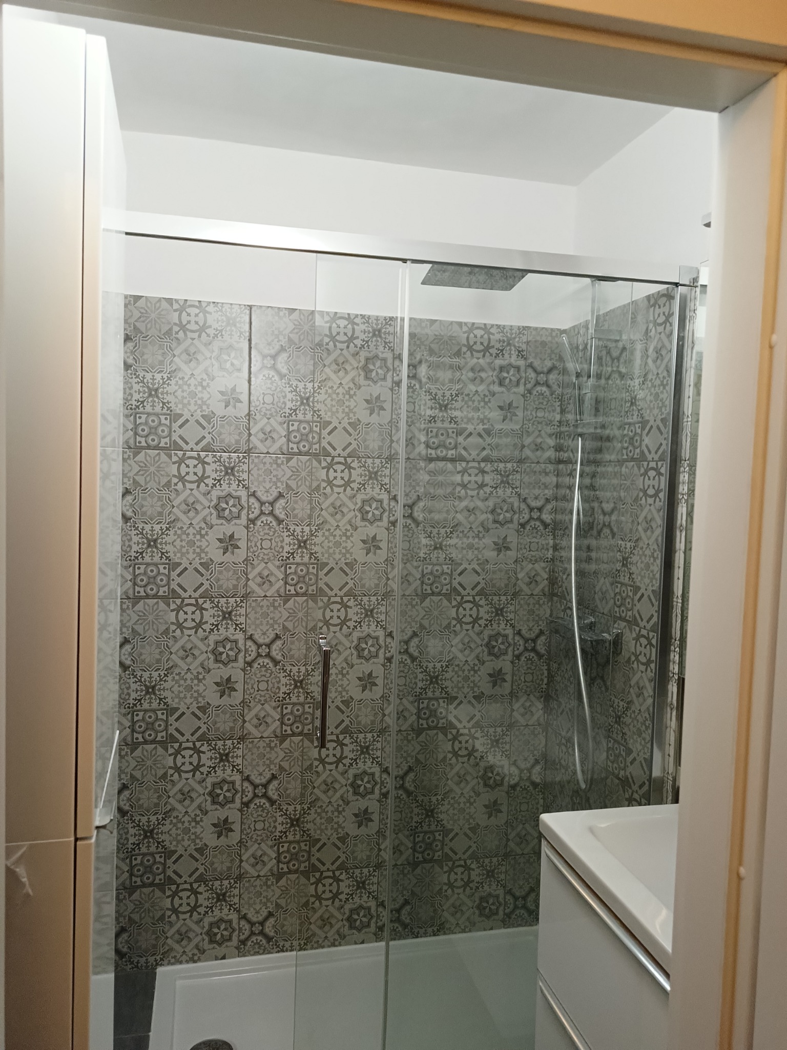 Nowoczesna kabina prysznicowa z szarą mozaiką, widok z zewnątrz, fragment białej szafki łazienkowej po prawej stronie.