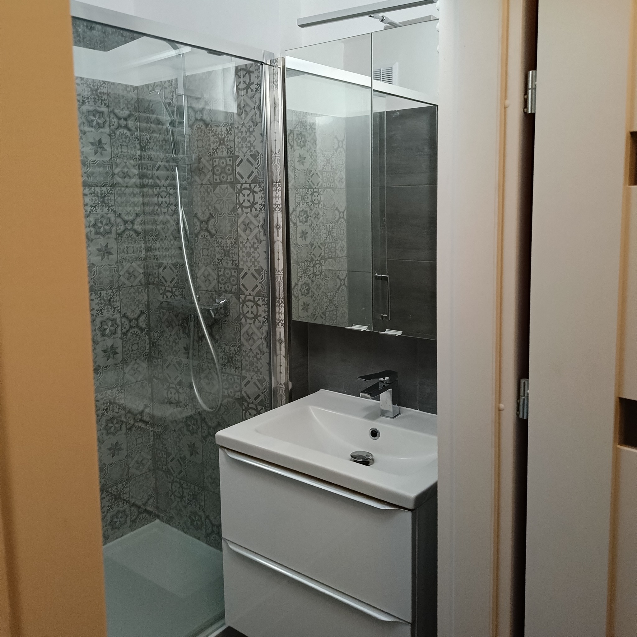 Nowoczesna łazienka z kabiną prysznicową wyłożoną mozaiką w odcieniach szarości, białą umywalką z szafką i lustrem z oświetleniem nad umywalką, widok zza otwartych drzwi.