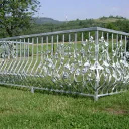 Surowa, metalowa balustrada balkonowa z dekoracyjnymi elementami roślinnymi, stojąca na trawie przed domem w słoneczny dzień.