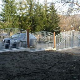 Kowalstwo Piotr Ficoń - Biała brama wjazdowa z drewnianymi słupkami i falistym wzorem, za którą widać zaparkowany samochód osobowy.