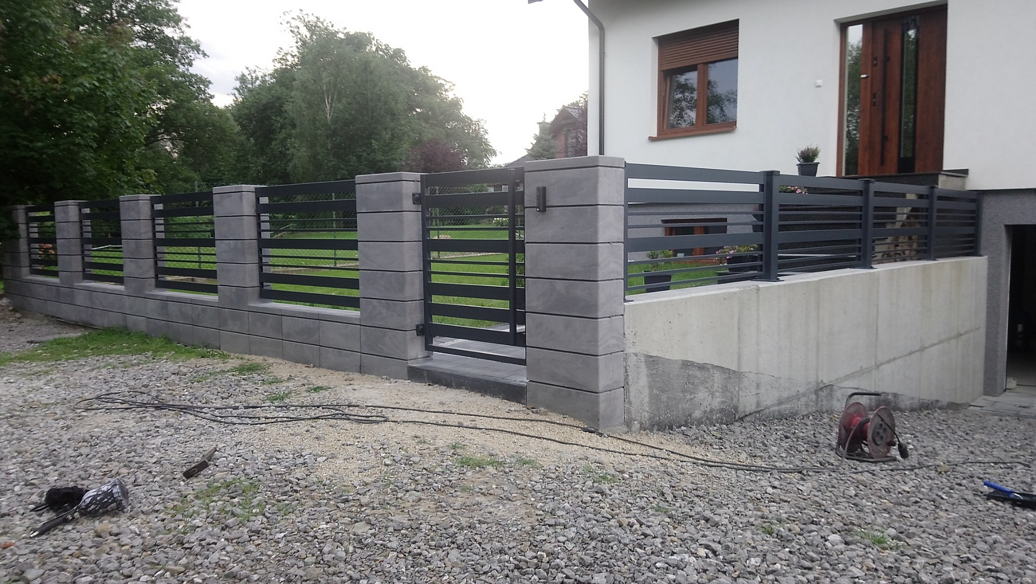 Nowoczesne ogrodzenie panelowe z bramą wjazdową, szare słupki z betonowych segmentów, poziome metalowe przęsła, widoczne narzędzia na żwirowym podłożu.