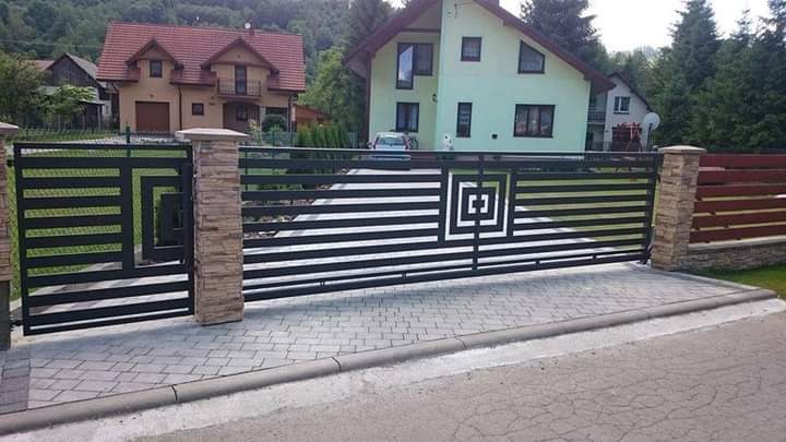 Nowoczesna, pozioma brama wjazdowa z czarnych, płaskich profili, z kwadratowym motywem dekoracyjnym, osadzona na słupkach obłożonych kamieniem elewacyjnym, na tle zabudowy jednorodzinnej.