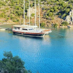 Luksusowy jacht żaglowy zacumowany przy pomoście w malowniczej zatoce z zielonymi wzgórzami w tle, w słoneczny dzień.