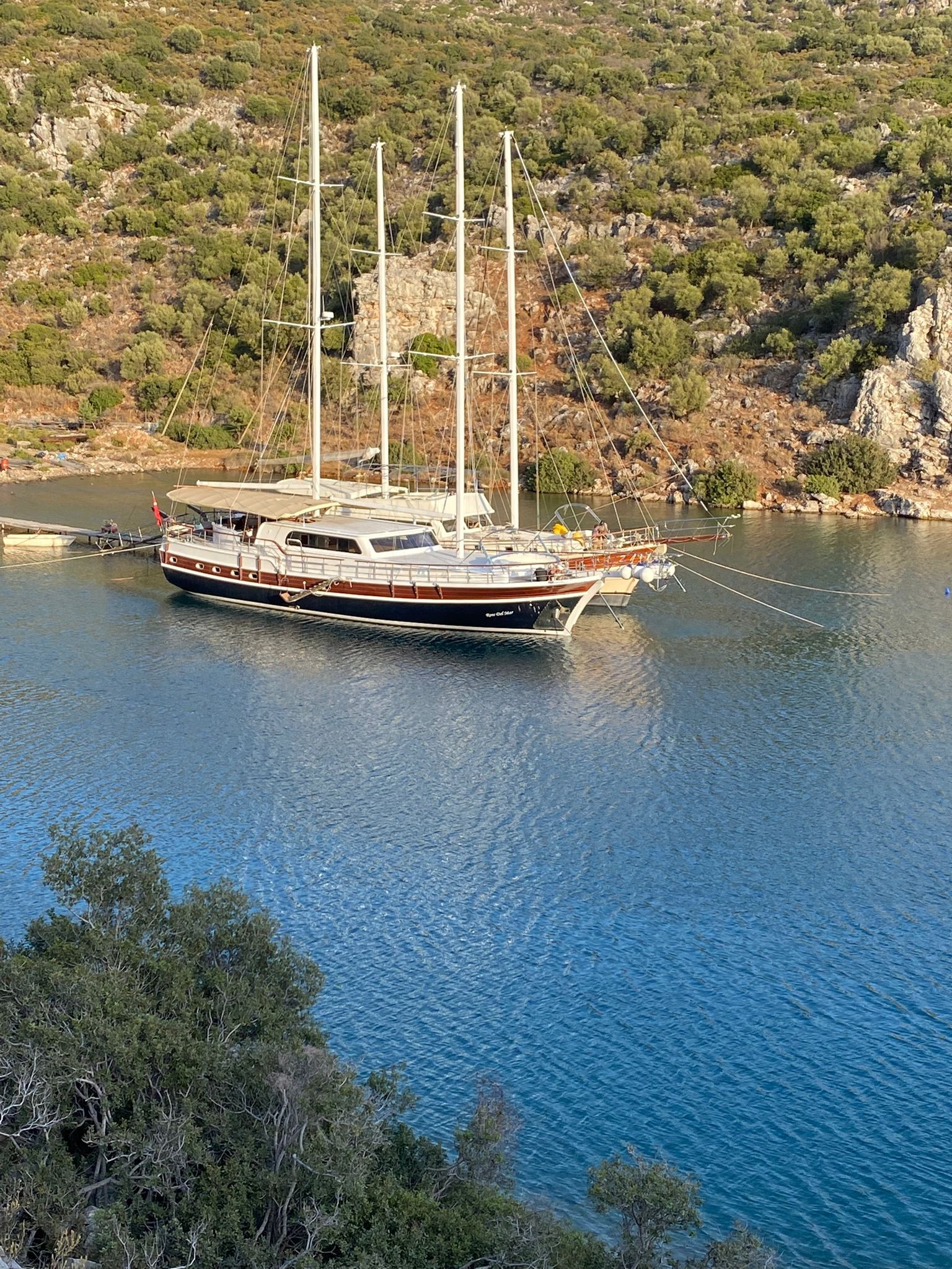 Elegancki jacht z trzema masztami zacumowany w spokojnej zatoce, otoczony zielonymi wzgórzami. Scena z wakacji w Warszawie, sugerująca luksusowe rejsy.