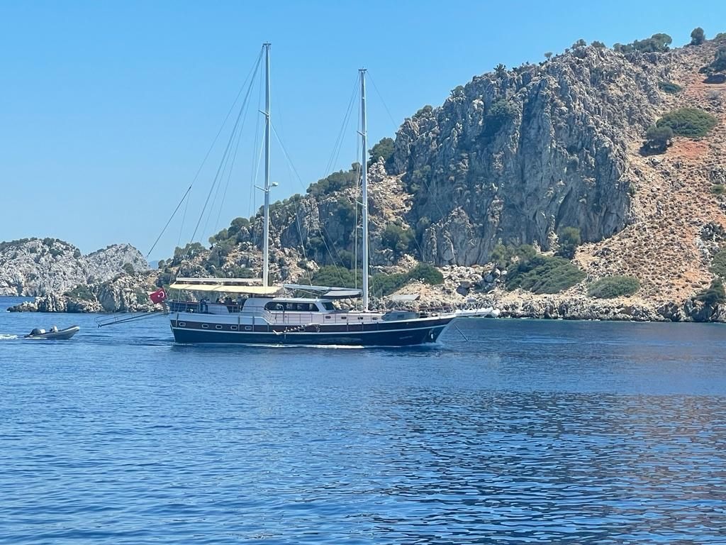Dwumasztowy jacht motorowy na tle skalistego wybrzeża z niewielką motorówką holowaną za rufą, błękitne morze i bezchmurne niebo.