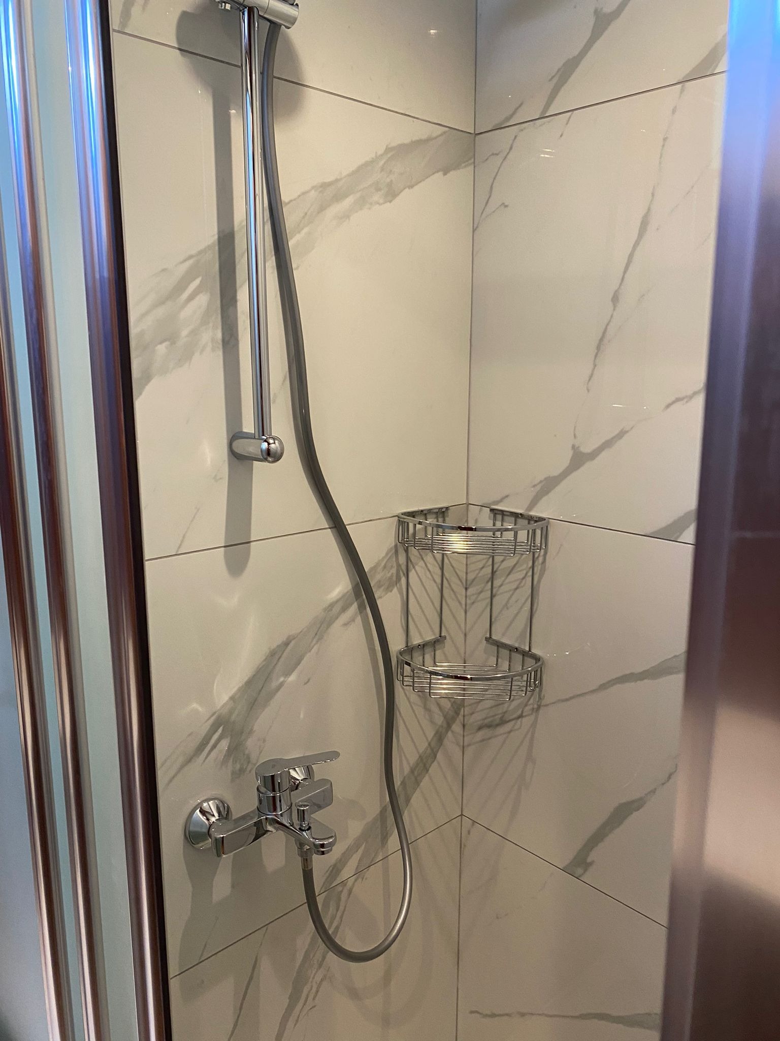Błyszcząca, chromowana bateria prysznicowa, słuchawka i półka narożna na marmurowej ścianie w kabinie prysznicowej z brązowymi profilami.
