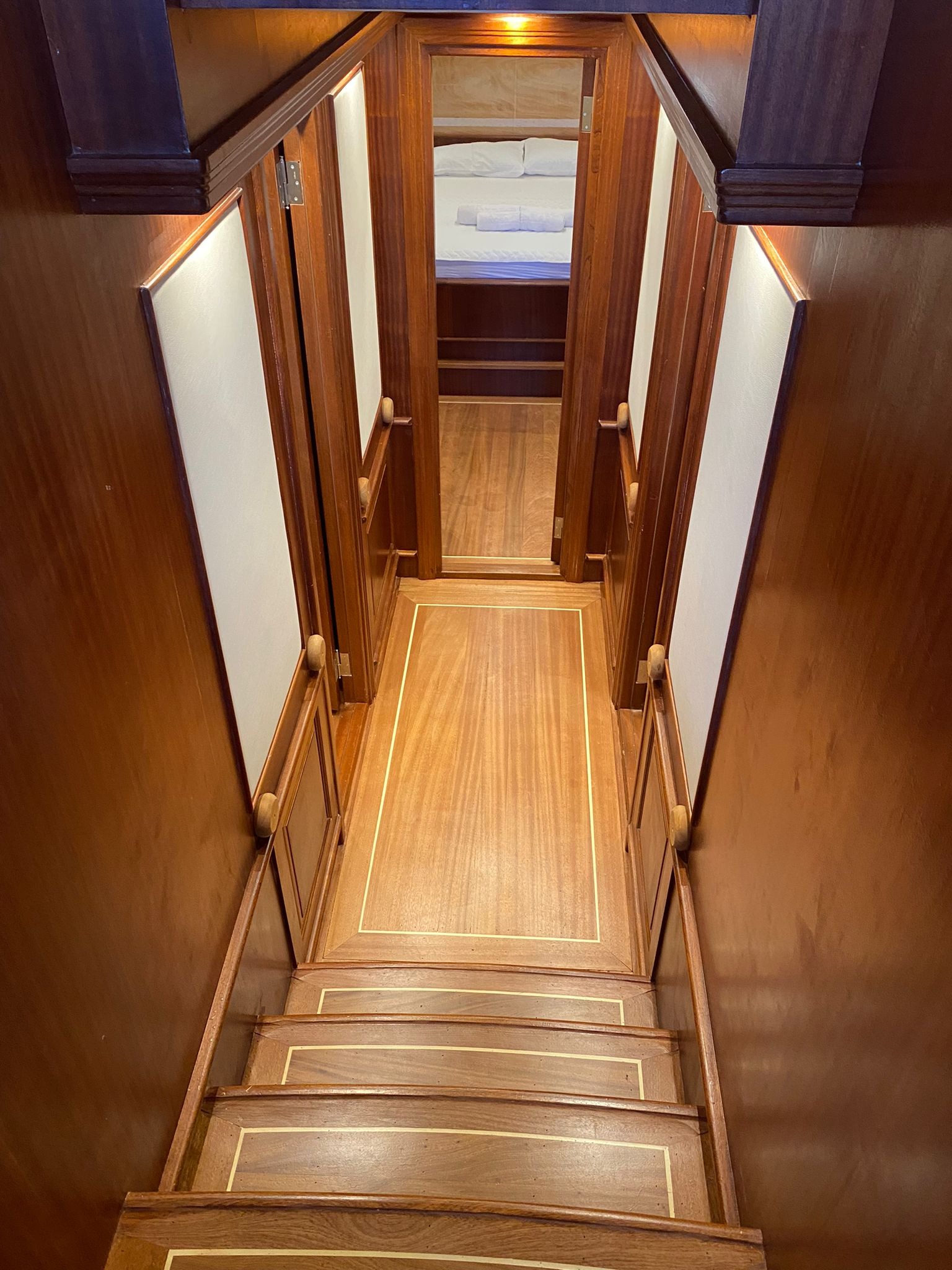 Wnętrze luksusowego jachtu: schody z ciemnego drewna z jasnymi intarsjami prowadzące do kajuty z pościelonym łóżkiem w tle.
