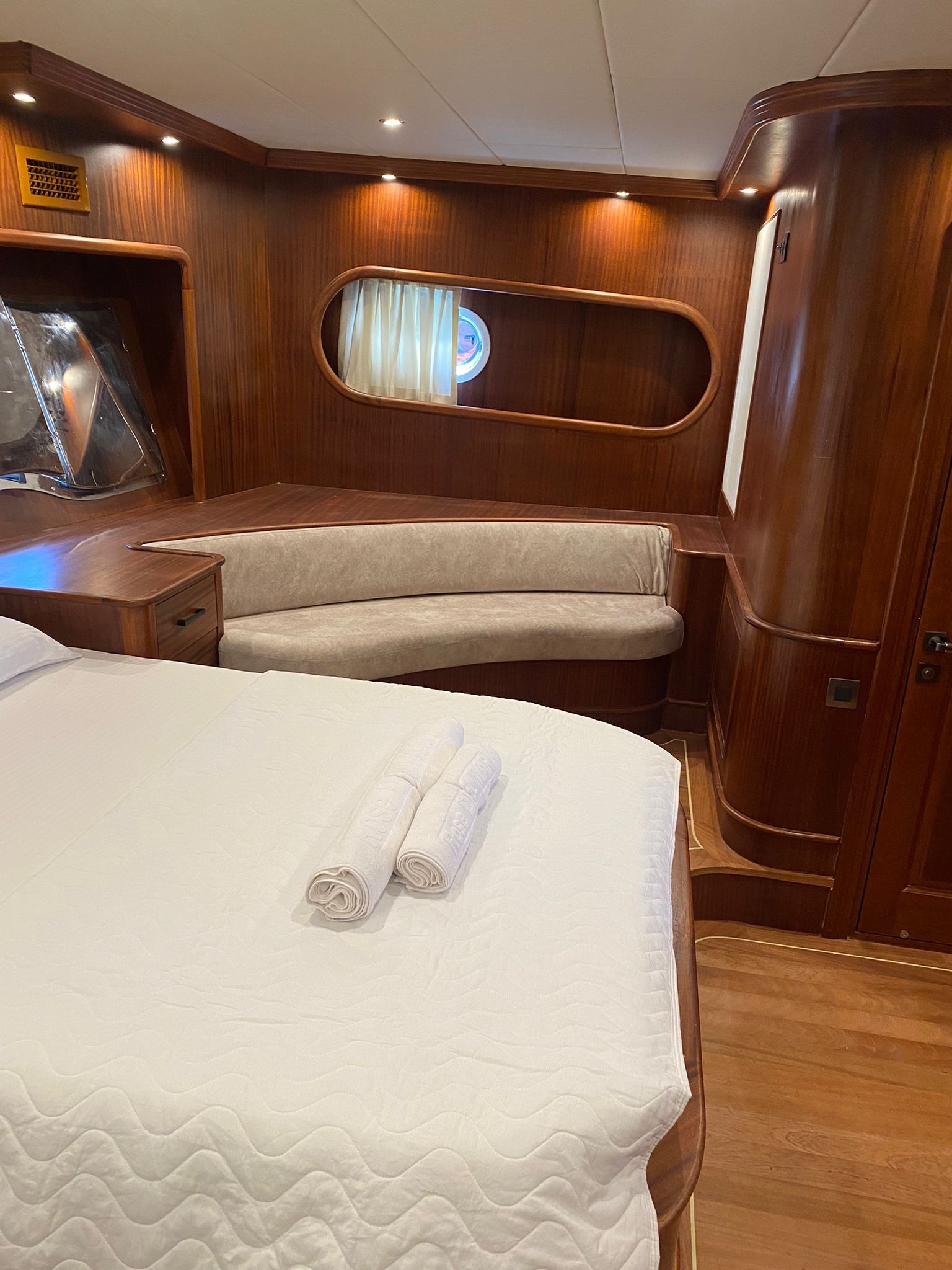 Kabinę jachtu z drewnianymi panelami, łóżkiem z białą pościelą i zwiniętymi ręcznikami oraz oknem w owalnej ramie.