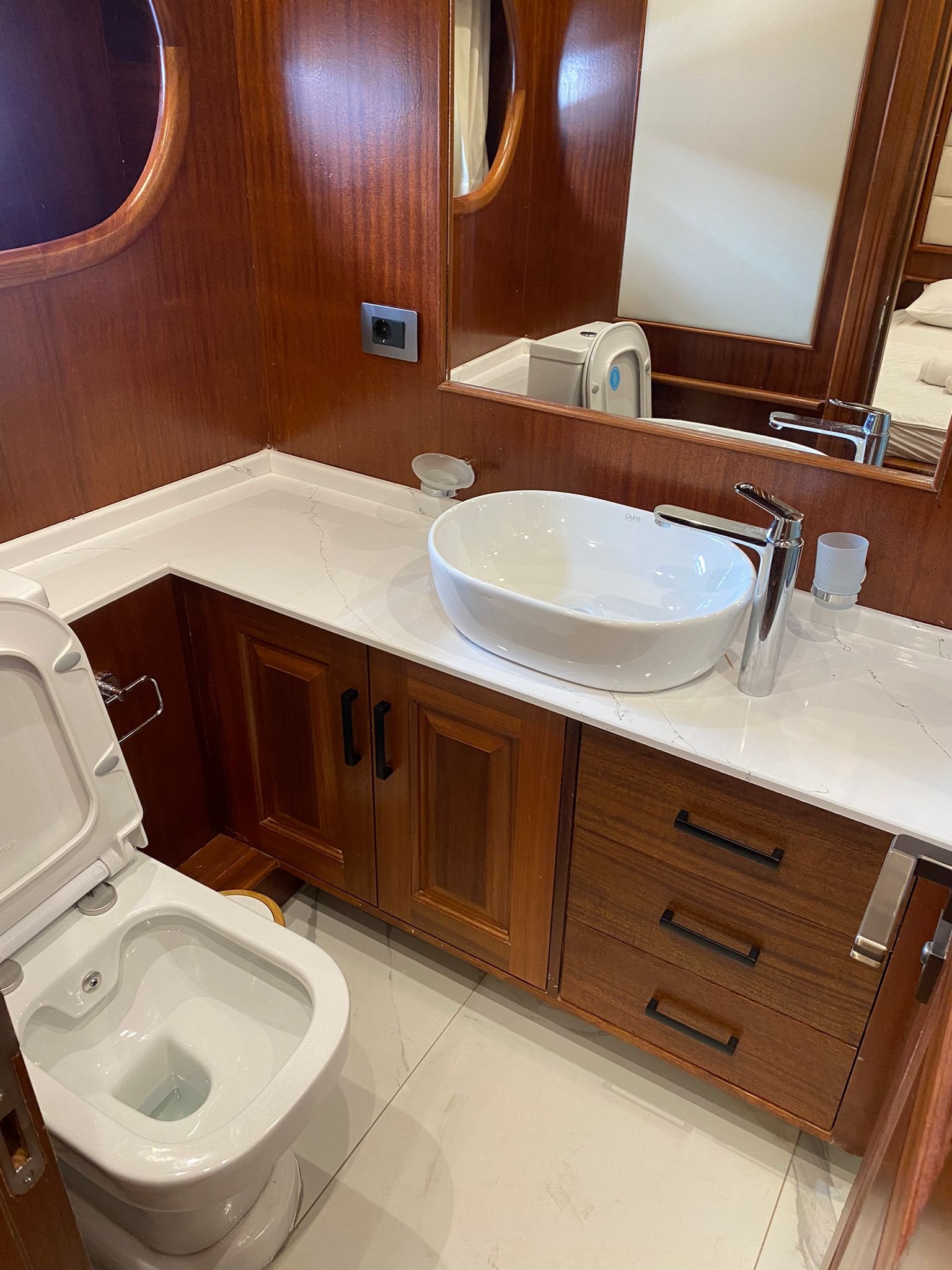 Wnętrze łazienki na jachcie z drewnianymi wykończeniami, białą umywalką nablatową i toaletą z otwartą klapą.