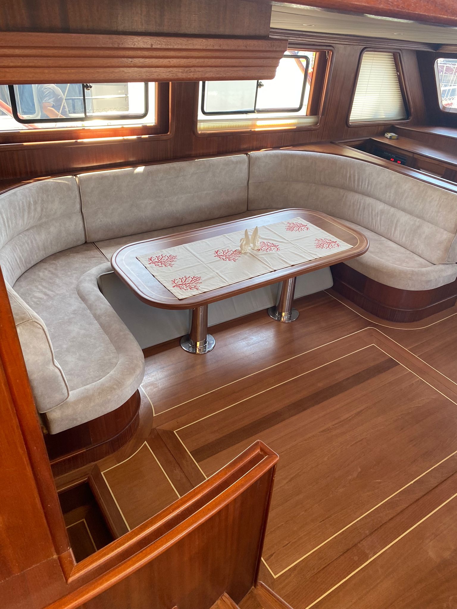 Wnętrze jachtu z drewnianą podłogą i obiciem, stół z dekoracyjnym obrusem w motywy koralowe, widoczne okna z roletami.