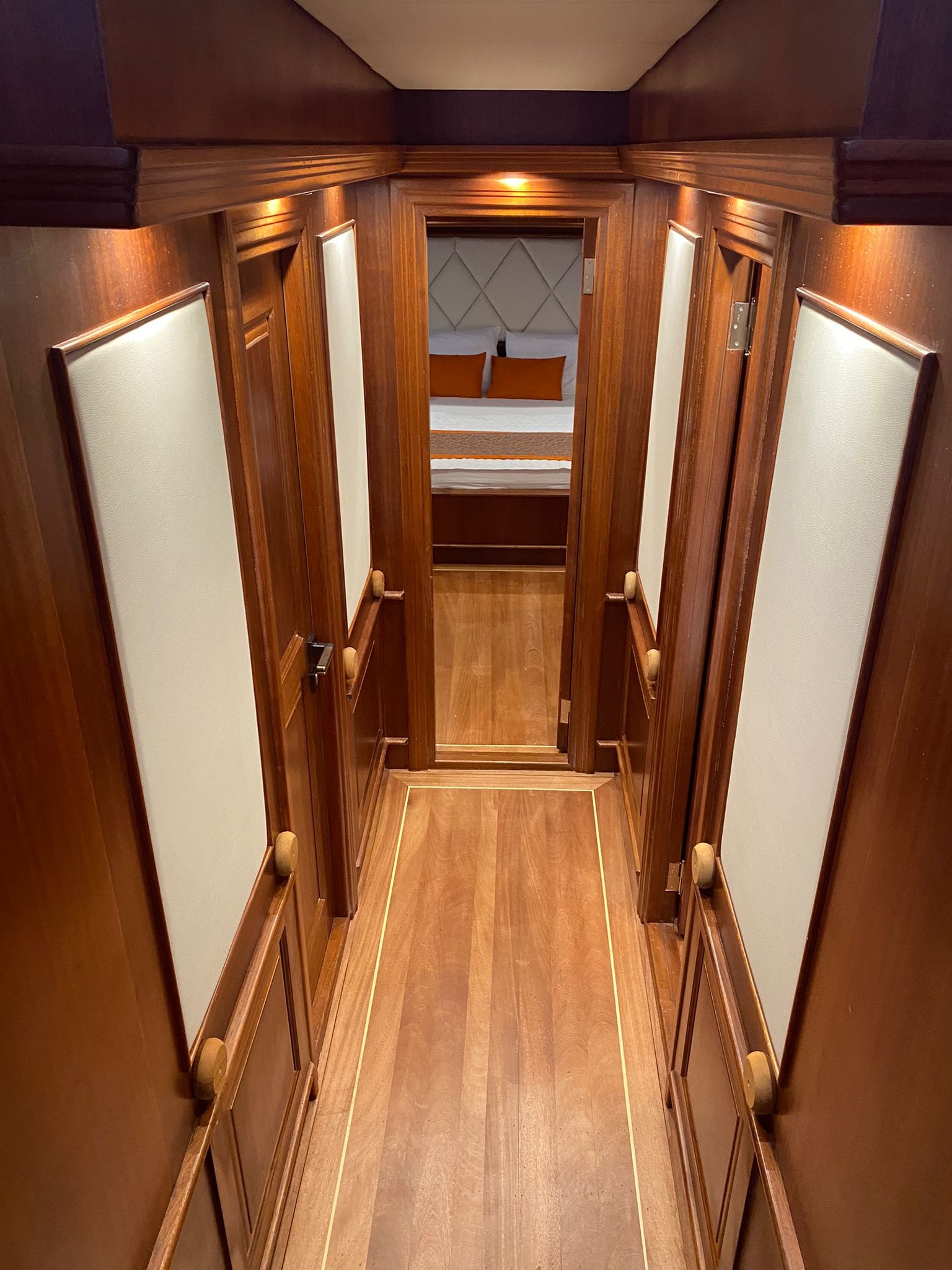 Wnętrze luksusowego jachtu: korytarz z drewnianą boazerią, kremowymi panelami i widokiem na sypialnię z pomarańczowymi poduszkami.