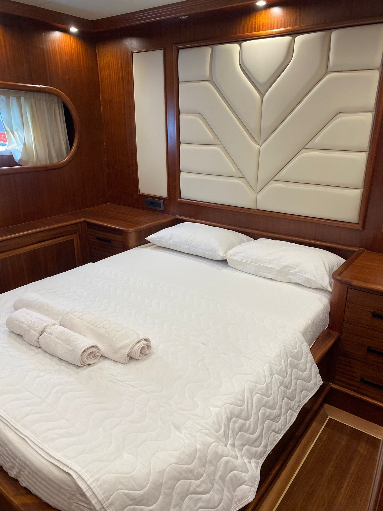 Wnętrze kabiny jachtu: biała pościel na łóżku, zwinięte ręczniki z napisem, drewniane wykończenie ścian i mebli, dekoracyjny zagłówek z jasnej tapicerki.