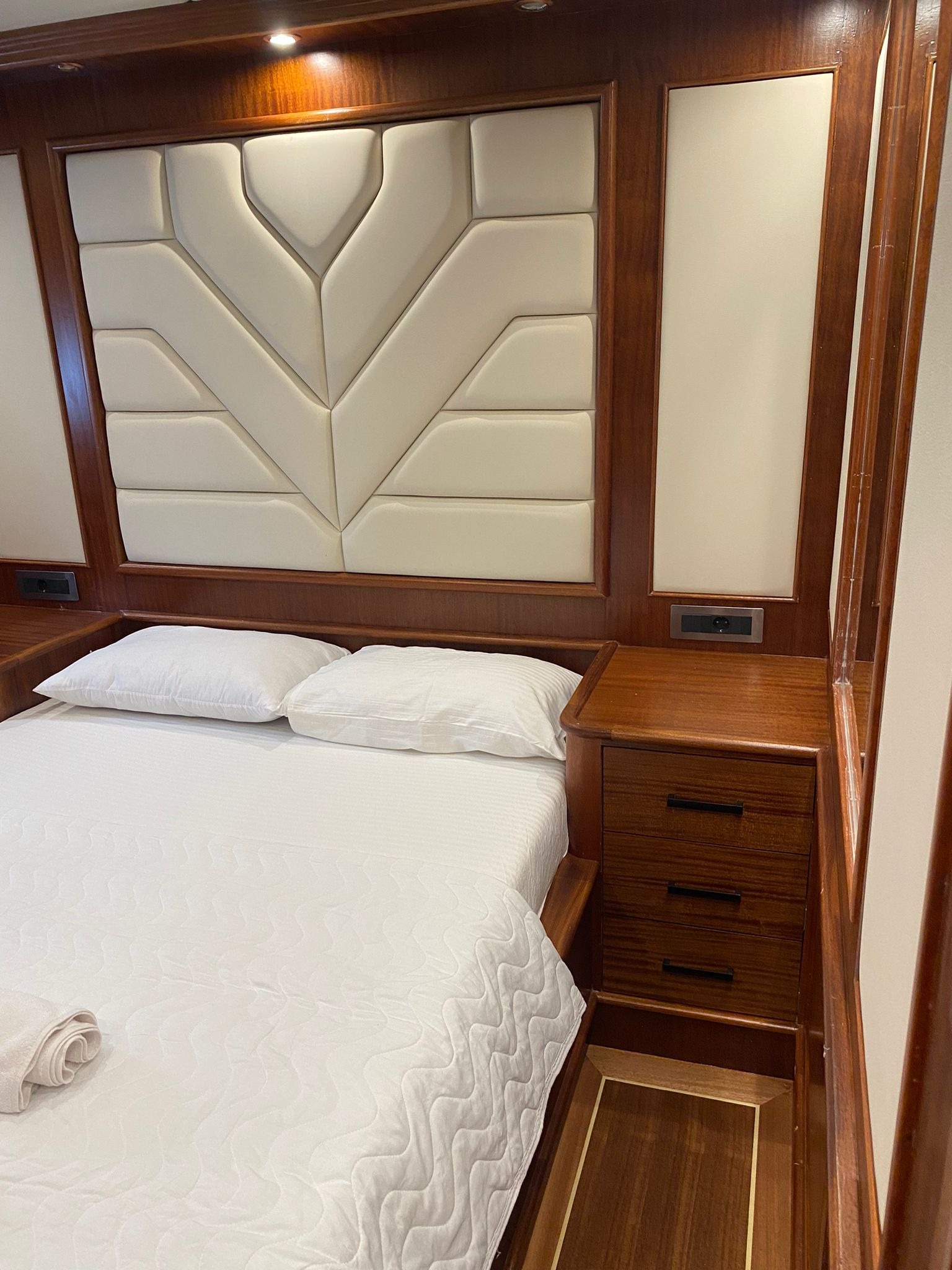 Kabina jachtu z łóżkiem, drewnianymi szafkami nocnymi i ozdobnym, kremowym zagłówkiem w kształcie liścia.