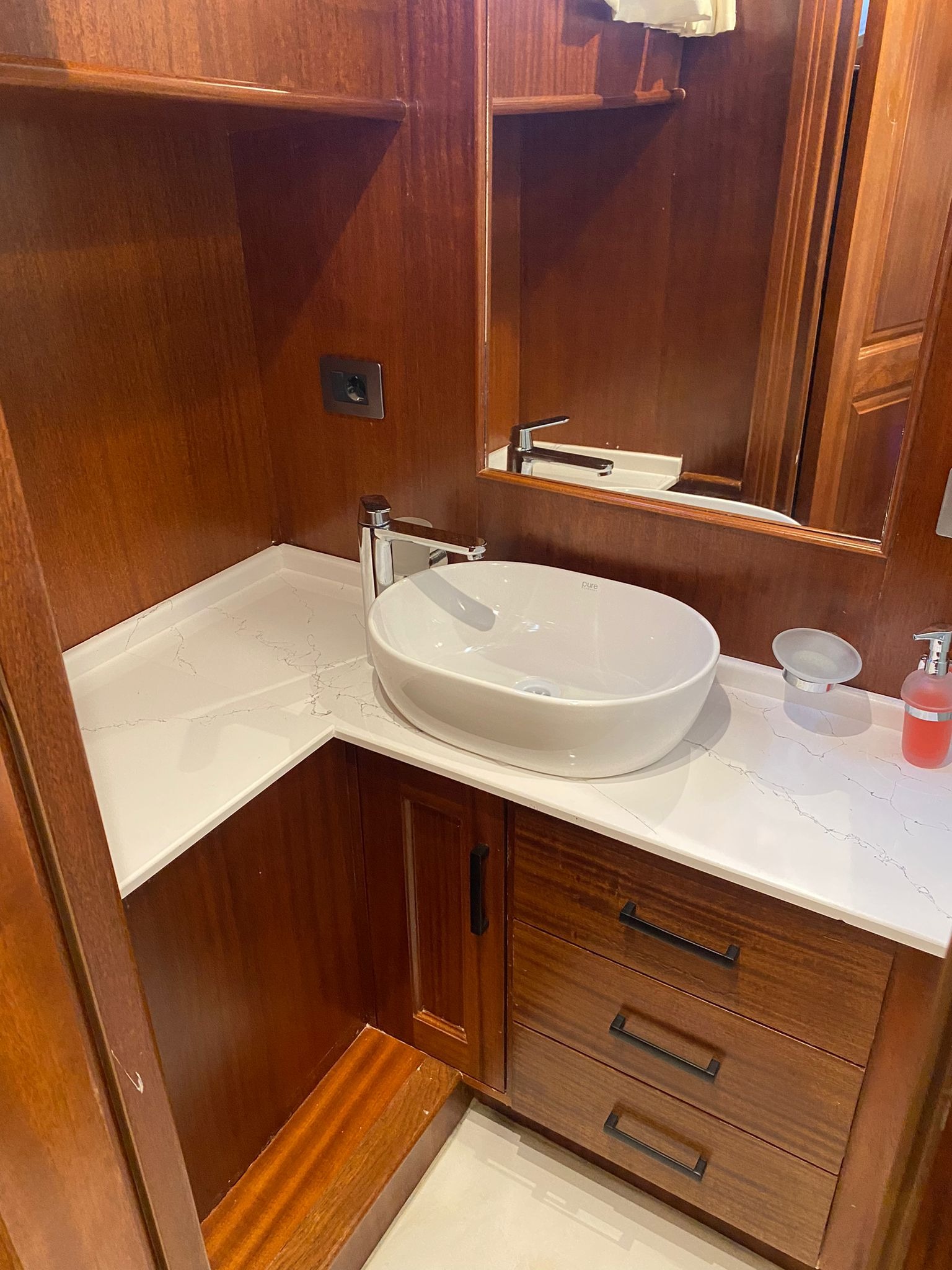 Wnętrze kabiny jachtu: umywalka z białej ceramiki na blacie z białego konglomeratu z brązowymi szafkami z drewna egzotycznego, chromowana bateria i dozownik mydła.