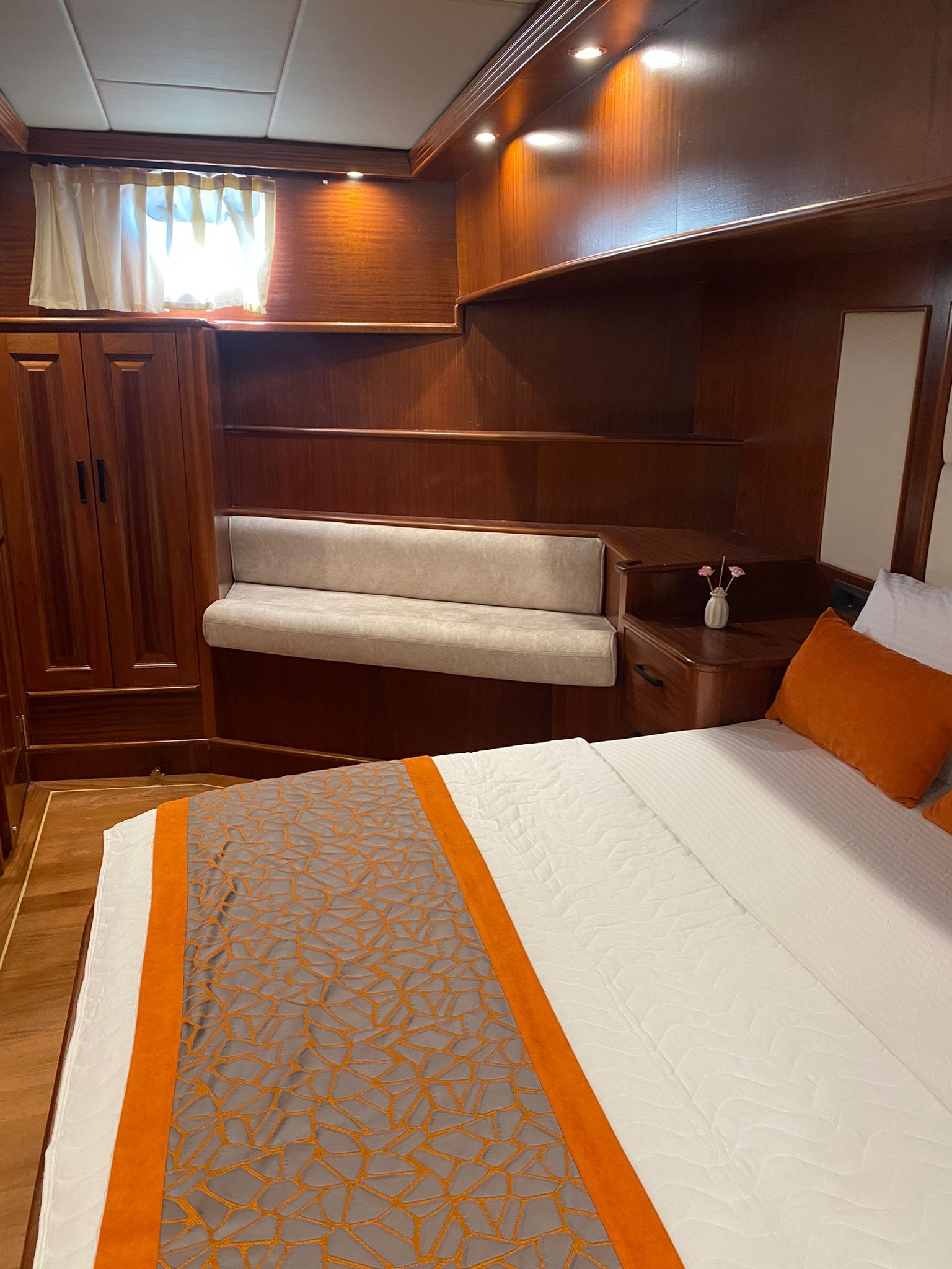 Wnętrze kabiny jachtu z drewnianą zabudową, łóżkiem z pomarańczowymi poduszkami i narzutą w geometryczne wzory, mała sofa pod oknem.