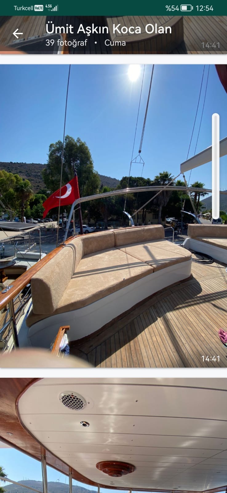 Wnętrze luksusowego jachtu z drewnianym pokładem, beżowymi materacami do opalania i turecką flagą powiewającą na wietrze, w tle zielone wzgórza i błękitne niebo.