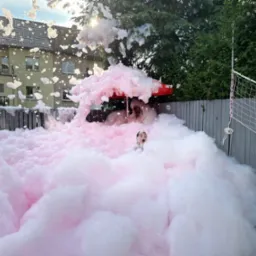 Dziewczynka z otwartymi ustami zanurzona w różowej pianie na imprezie plenerowej, widoczny fragment czerwonego parasola i siatka do badmintona.