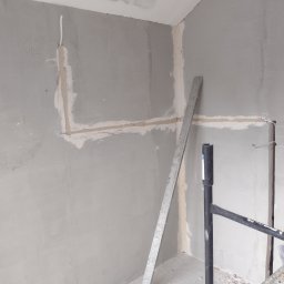 Eryk Jabłoński wykończenia dekarstwo brukarstwo i gwarancja - Ściana w trakcie prac przygotowawczych do wykończenia, widoczne ślady po instalacjach elektrycznych, częściowo wypełnione bruzdy, pionowa poziomica oparta o ścianę, fragment rusztowania.