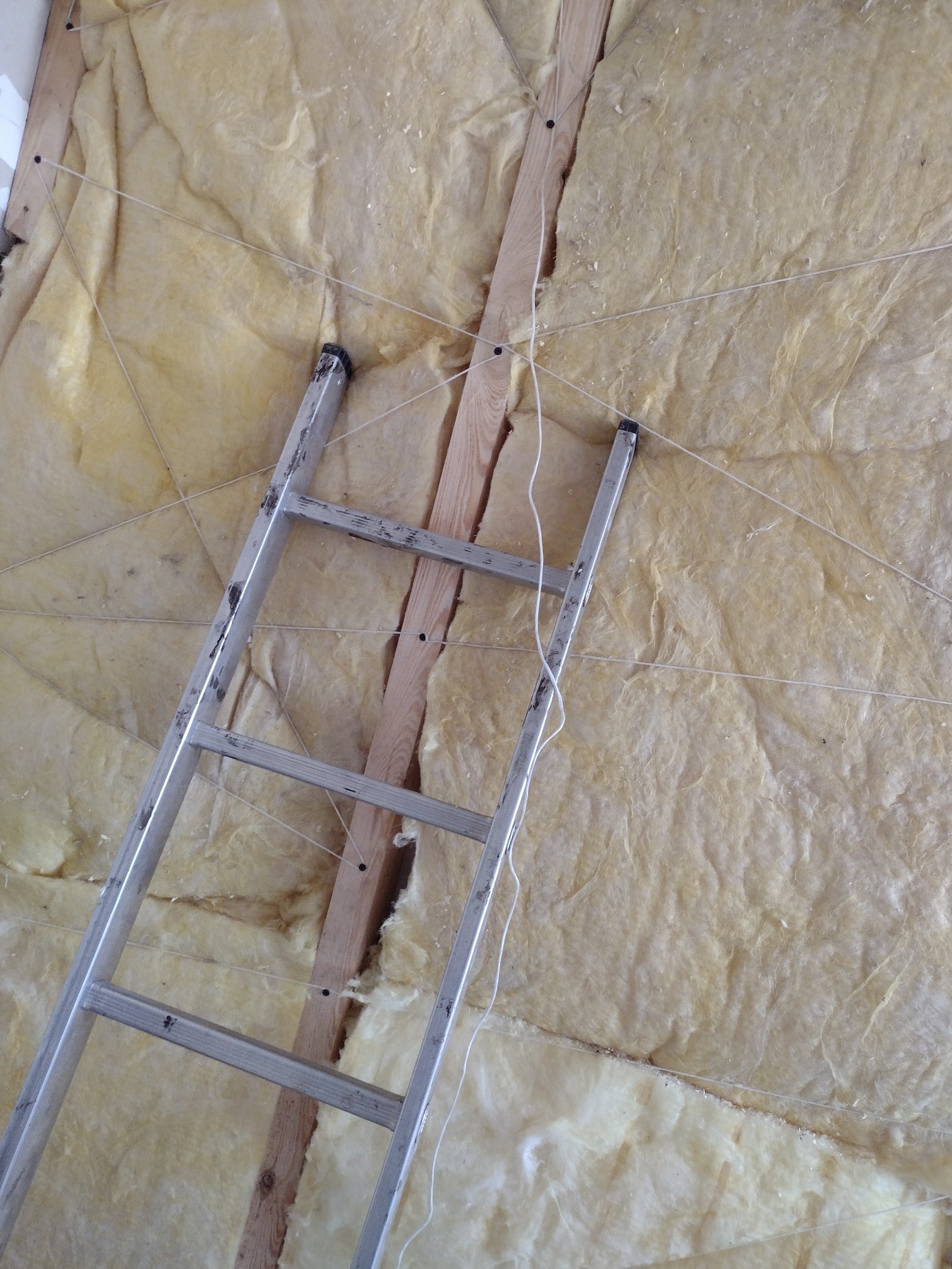 Montaż izolacji z wełny mineralnej na poddaszu, widoczna aluminiowa drabina i sznurki mocujące do drewnianej konstrukcji.