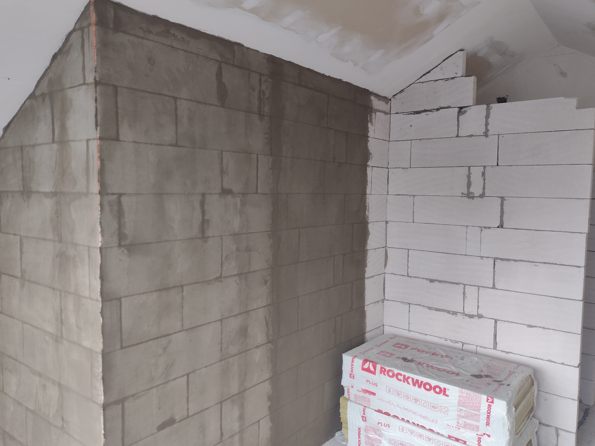Ściana z bloczków częściowo pokryta świeżą warstwą szarej zaprawy, widoczny laserowy wskaźnik pionu oraz paczki wełny mineralnej ROCKWOOL u podstawy ściany.